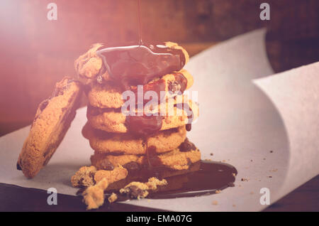 In salsa di cioccolato colata su una pila di biscotti al cioccolato su bianco arricciatura carta da forno contro un legno scuro sfondo, wit Foto Stock