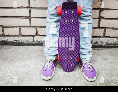 Adolescente in jeans e gumshoes detiene viola skateboard vicino dal grigio urbano parete di mattoni Foto Stock