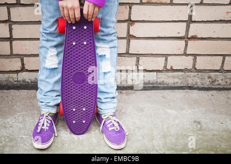 Ragazza adolescente in jeans e gumshoes detiene skateboard vicino dal grigio urbano parete di mattoni Foto Stock