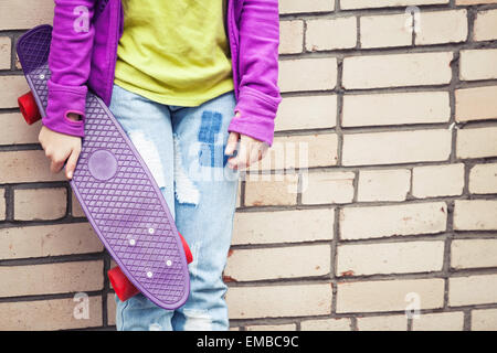 La bionda ragazza adolescente in jeans e colorato i vestiti sportivi skateboard tiene vicino dal grigio urbano parete di mattoni Foto Stock