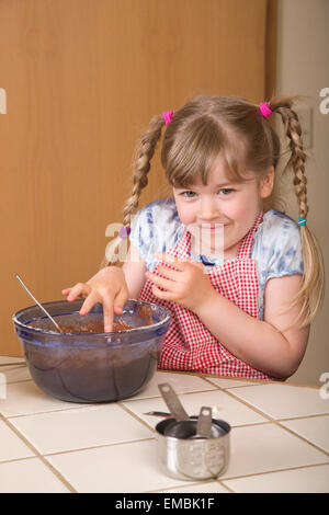 Bambina di cinque anni di intrufolarsi un morso di impasto dal recipiente di miscelazione di bundt cioccolato torte Foto Stock
