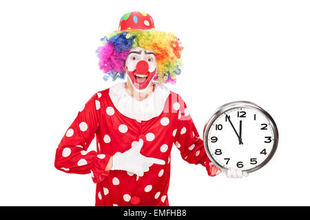 Giovane maschio clown tenendo un grande orologio da parete in una mano e rivolto verso l'orologio con gli altri isolati su sfondo bianco Foto Stock