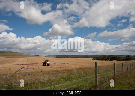 Un agricoltore di colture di spruzzatura in un campo di grano con reticolati di filo spinato sotto un cielo nuvoloso Foto Stock