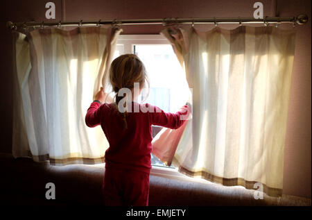 Una sonnolenta giovane ragazza in pigiama apre le tende a guardare fuori dalla finestra all'inizio di un nuovo giorno Foto Stock