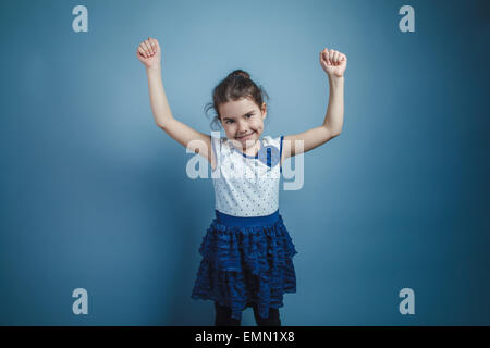 Una bambina di sette anni aspetto europeo brunette sollevate le braccia sorridente su sfondo grigio, gioia Foto Stock