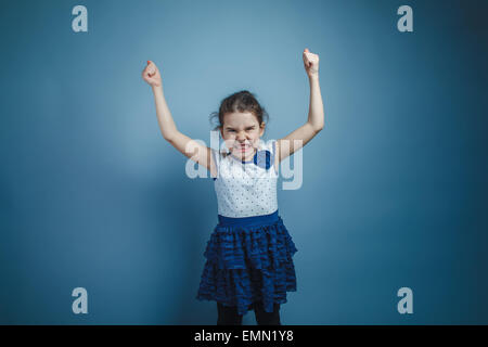 Una bambina di sette anni aspetto europeo bruna ha sollevato le sue mani fino arrabbiato su sfondo grigio, odio Foto Stock