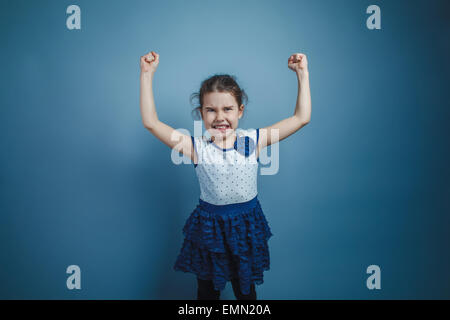 Una bambina di sette anni aspetto europeo brunette mostra i muscoli delle mani su sfondo grigio, la forza Foto Stock