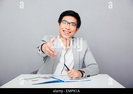 Sorridente imprenditore asiatici seduti a tavola e stretching mano per stretta di mano su sfondo grigio. Guardando la fotocamera Foto Stock