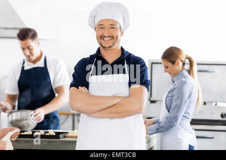 Fiducioso chef in piedi con le braccia incrociate mentre i colleghi che lavorano in Foto Stock