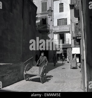 Degli anni Cinquanta, storica strada laterale, Palma di Maiorca, Spagna, uomo di lavoro spingendo un vuoto carrello di legno. Foto Stock