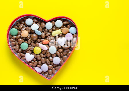 A forma di cuore scatola riempita con piccoli cioccolatini sfere con un sacco di loro sparse su sfondo giallo Foto Stock