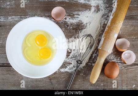 La cottura di sfondo con uova, uova crude in un piatto da portata, gusci d'uovo, la farina, il mattarello e frusta su uno sfondo di legno. Vista dall'alto. Foto Stock