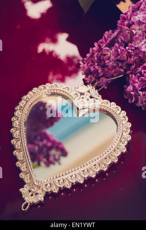 Elegante a forma di cuore specchio con cornice in argento e fiori lilla sulla superficie rossa. Focus sul mirror. Foto Stock