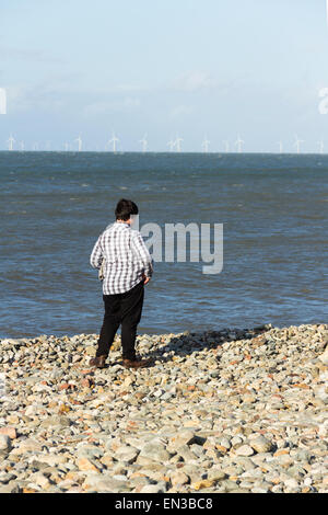 Un ragazzo adolescente permanente sulla spiaggia di ciottoli a Llandudno, il Galles del Nord guardando le turbine eoliche del Rhyl Appartamenti fattoria eolica. Foto Stock