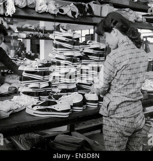 Fine 1950s, storico, una lavoratrice cinese femminile in piedi ad un banco da lavoro mettendo i lacci in tessuto sintetico, le sneakers con suola in gomma o scarpe tipo trainer, Hong Kong. Note anche come décolleté, queste scarpe leggere sono state prodotte in grandi quantità e sono diventate popolari per il tempo libero o casual abbigliamento e sport. Foto Stock