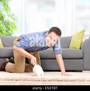 Giovane uomo aspira un tappeto con un palmare aspirapolvere e sorridente a casa Foto Stock