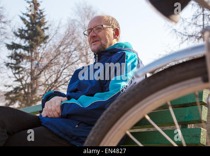 Ritratto di eldely uomo che indossa gli occhiali, pantaloni neri e una giacca blu seduta su una panchina vicino la sua bicicletta in un parco della città Foto Stock
