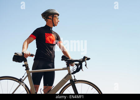 Immagine di determinata ciclista maschio che guarda lontano mentre in piedi contro il cielo. Montare l'uomo di indossare abbigliamento sportivo e ciclismo casco cercando Foto Stock