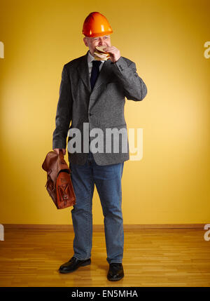 Ingegnere maschio in camera, mangia il panino e detiene vecchia valigetta in pelle, pavimenti in parquet e di parete di colore giallo Foto Stock