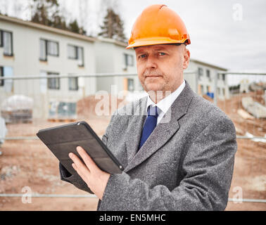 Ingegnere maschio utilizza computer tablet, egli indossare la tuta e l'elmetto, casa sito in costruzione sullo sfondo Foto Stock
