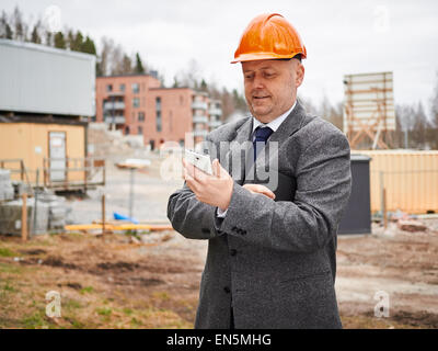 Ingegnere maschio utilizza il telefono cellulare, egli indossare la tuta e l'elmetto, casa sito in costruzione sullo sfondo Foto Stock