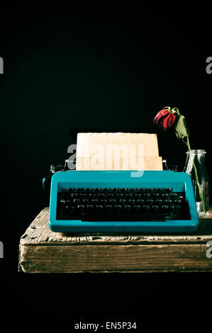 Una vecchia macchina da scrivere blu con una pagina vuota nel suo rullo e un appassì rosa rossa in un vaso di vetro su una tavola in legno rustico, contro Foto Stock