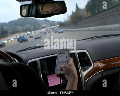 Scrivere la guida usa il driver femminile in l/h guidare il veicolo texting con iPhone smartphone durante la guida su occupato Interstate Highway California USA Foto Stock