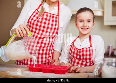 Adorabile ragazza guardando la fotocamera mentre sua madre il riempimento in forme di muffin con impasto liquido Foto Stock
