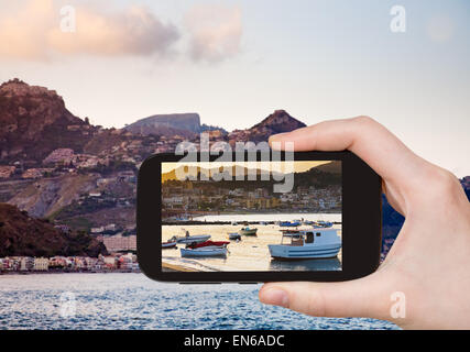 Concetto di viaggio - tourist prende foto di barche al tramonto, vicino alla città di Taormina, Sicilia, Italia sullo smartphone Foto Stock