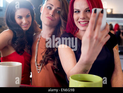 Tre giovani donne eleganti caffè al bar alla ricerca e ridere mentre prendendo selfie con il cellulare Foto Stock