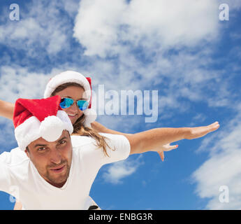 Sorridenti giovane piggyback insieme con le braccia aperte in rosso cappelli di Babbo Natale su blu cielo tropicale sfondo Foto Stock