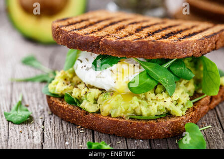 Sandwich alla griglia con avocado, uovo in camicia e rucola sul tavolo di legno Foto Stock