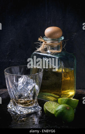 Bicchiere di tequila anejo con cubetti di ghiaccio e una bottiglia di tequila, servito con spicchi di lime su sfondo nero Foto Stock