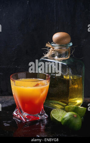 Bicchiere di tequila sunrise cocktail serviti con una bottiglia di tequila anejo affettato limes su sfondo nero. Rendendo la tequila sunris Foto Stock