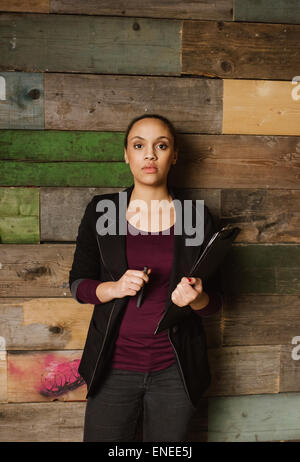 Ritratto di determinata giovane donna tenendo un clipboard in piedi contro una parete in legno guardando la fotocamera. African business donna Foto Stock