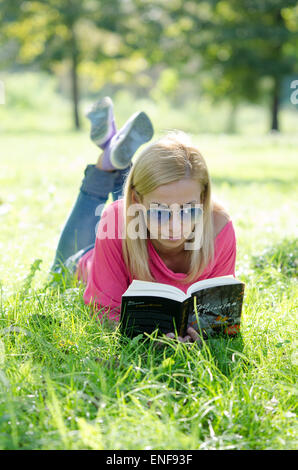 Giovane donna in jeans blu con occhiali da sole sdraiati sull'erba e libro di lettura Foto Stock