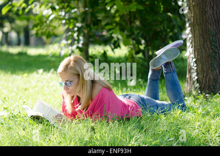 Giovane donna in jeans blu con occhiali da sole sdraiati sull'erba e libro di lettura Foto Stock