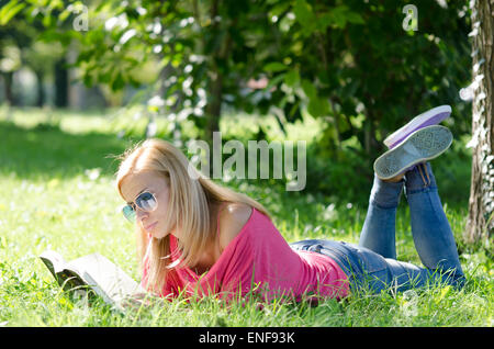 Giovane donna in jeans blu con occhiali da sole sdraiati sull'erba e libro di lettura Foto Stock