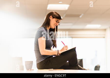 Una giovane donna seduta sulla sua scrivania la scrittura in un notebook con una penna rossa. Foto Stock