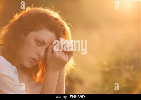 Giovane donna con capelli auburn posa in un campo illuminate dal sole al tramonto Foto Stock