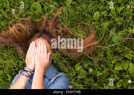 Giovane ragazza carina con tousled lunghi capelli sdraiati sull'erba verde che copre il viso con le mani. Foto Stock