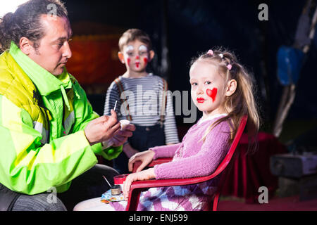 Ragazza bionda seduto in poltrona guardando verso la telecamera e avente compongono applicato alla faccia da uomo indossando giacca luminose con ragazzo Clown Foto Stock