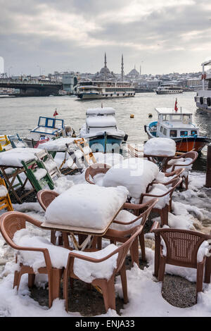 Coperta di neve cafe sedie sul lungomare di Karakoy, guardando attraverso il Golden Horn verso la moschea Yeni a Eminonu e Istanb Foto Stock