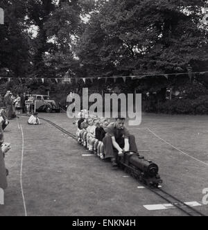 Degli anni Cinquanta, storico, padre con la sigaretta in bocca sterza il su piccola scala e a livello del suolo con modello di treno o di locomotiva in miniatura con bambini come passeggeri su un campo. Foto Stock