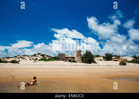 LAMU, Oceano Indiano, Kenya, Africa. Una giovane donna in un bikini si trova in acque poco profonde su una spiaggia con un vecchio castello-come edificio di distanza. Foto Stock