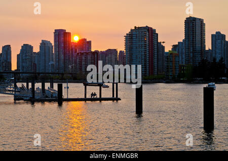 Bel tramonto su False Creek in Vancouver, con gli edifici della città in silhouette. Due persone sedute su una panchina sul molo Foto Stock