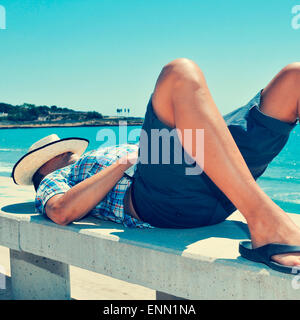 Un giovane uomo caucasico con un cappello di paglia sul suo volto disteso in un banco di strada vicino al mare Foto Stock