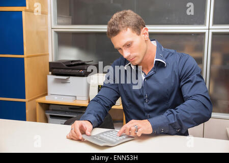 Bel giovane uomo che lavora in ufficio alla sua scrivania utilizzando la calcolatrice Foto Stock