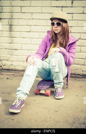 Bella ragazza adolescente nel cappello e occhiali da sole si siede su uno skateboard urbano vicino a un muro di mattoni, verticale Foto Stock