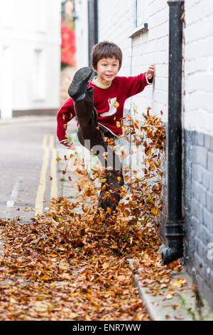 Bambino caucasico, ragazzo, 8-9 anni, camminando lungo una strada coperta di uscita verso lo spettatore, calciando le foglie in alto in aria. Autunno. Foto Stock